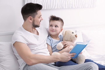 Dad and son reading interesting book while sitting on bed at home
