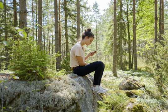 Sweden, Ostergotland, Girl (14-15) sitting on rock in forest using smartphone