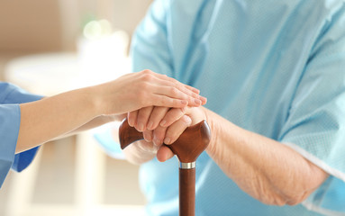 Young doctor holding hands of elderly woman on light background, closeup