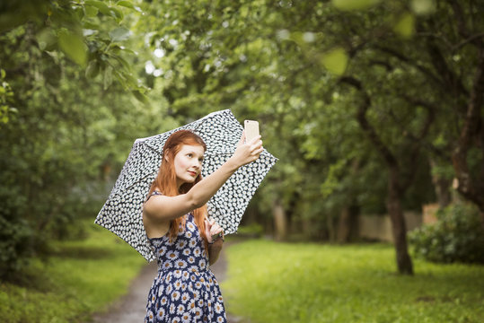 Finland, Pirkanmaa, Tampere, Woman wearing floral dress standing with umbrella in park and taking selfie