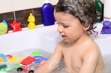 Beautiful toddler taking a bath in a bathtub with bubbles. Cute kid washing his hair with shampoo in the shower and splashing water everywhere. Boy playing with toys in the tub