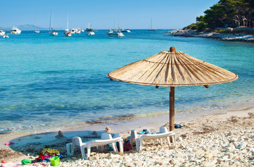 Fototapeta premium One yellow straw umbrella and two chairs on the pebble sandy beach. Many yachts in the azure blue sea lagoon in a bay near the coast of an island. Blue cloudless sky. Dugi otok, Croatia
