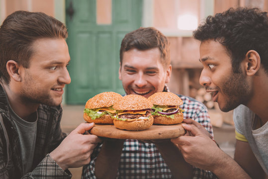 Excited young men looking at fresh tasty hamburgers on wooden board