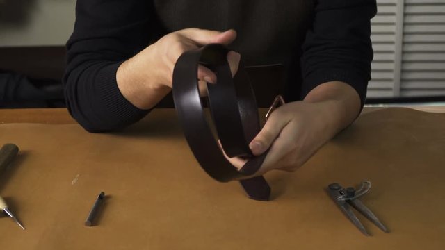 Smiling young man inspecting a leather belt he made at the tabel. Making a leather product. 
