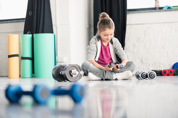 Cute smiling little girl sitting on floor and exercising with dumbbells in gym