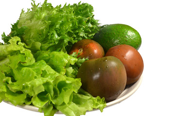 Fresh washed vegetables to prepare a green salad on large ceramic dish: ripe avocado, three black tomatoes 