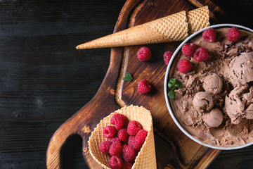 Bowl of homemade chocolate ice cream with fresh raspberries, mint and waffle cone on wood serving board over black burnt wooden background. Top view with space