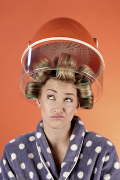 A woman looks dissatisfied with hair curlers in her hair sitting under the hood of a hair dryer, dressed in a bathrobe
