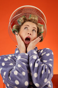 A shocked woman in a bathrobe with hair curlers in her hair sits under a hair dryer hood