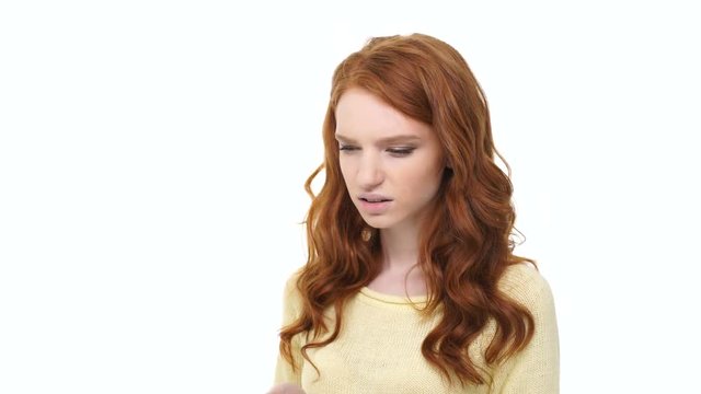 Young displeased annoyed woman with long red hair typing message on smartphone over white background