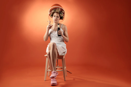 Woman with hair rollers sitting under drying hood with beer and cigarette in her hands