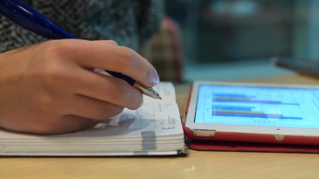 Close-up shot of a woman taking notes in notebook looking at tablet computer with information on business