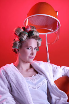Eine Frau mit Lockenwickler im Haar unter Trockenhaube in Dessous und Bademantel gekleidet