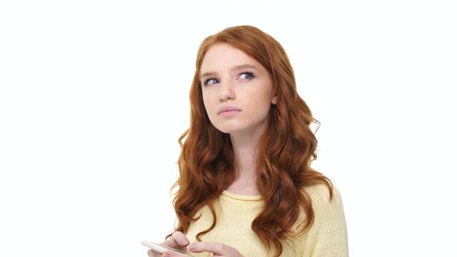 Pensive young woman with red long curly hair texting message on mobile phone isolated over white