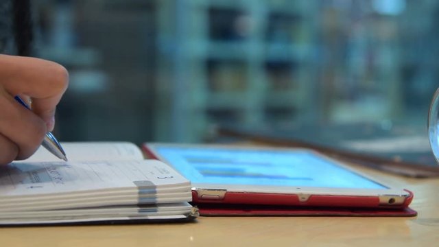 Close-up shot of a woman taking notes in notebook using touch pad with business information
