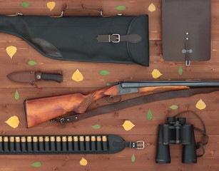 The gun, binoculars,bag, knife, leather bandolier and case for guns on a wooden background. Flat lay conception. Decorated with dried leaves. Hunting season. Top view. - 143416551