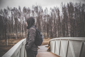Fototapeta premium Sad, lonely young man on the footbridge in the cold, rainy spring day. Gloomy atmosphere.