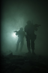 Fototapeta na wymiar Black silhouettes of pair of soldiers in the smoke haze moving in battle operation. Back light