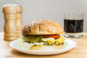 Vegetarian burger served on a white plate. Vegetarian hamburger sandwich with falafel cheak-peas cutlet, fresh vegetables and glass of soft cola drink on wood kitchen table