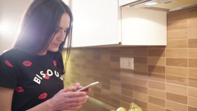 Young beautiful woman in home clothes is talking with a friend on the telephone and cooking a breakfast at home. She adds a yellow pepper in the pan and makes a photos of the food on her smartphone.