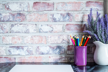 Colored pencils in a mug, vase of lavender flowers, white paper on a table against a brick vintage wall.