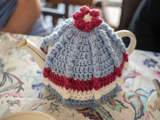 Vintage style teapot and knitted wool tea cosy or cozy on a patterned table cloth with a lace mat next to it.