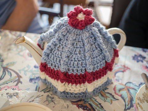 Vintage Style Teapot And Knitted Wool Tea Cosy Or Cozy On A Patterned Table Cloth With A Lace Mat Next To It.