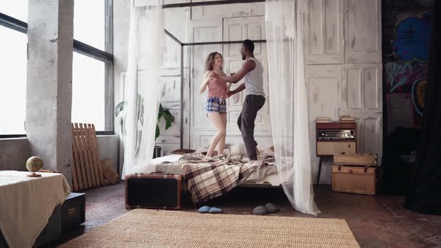 Young multiethnic couple in pajamas dancing, jumping on the bed. African boy and europen woman have fun in the morning.