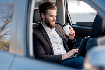 Handsome businessman sitting with coffee to go on the backseat of the car
