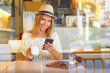 Cafe with wifi. Young pretty woman in hat using smartphone while drinking coffee.