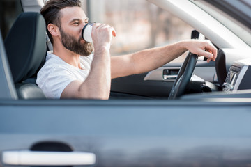 Handsome man dressed cassual in white t-shirt driving a car with coffee to go
