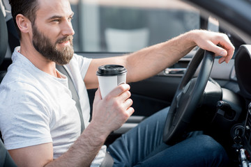 Handsome man dressed cassual in white t-shirt driving a car with coffee to go