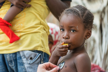 Stethoscope exam of African little girl eating fruit.Unrecognizable people in the background