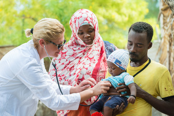 Female Caucasian doctor listening breath and heart beat of little African baby with stethoscope.Father holding the baby, mother looking at baby