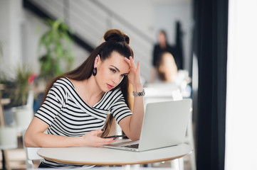 Fototapeta premium Woman sitting in a cafe in front of a laptop thinking about a problem with her hands touching head. Adult lady trying to find solution.