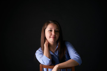Portrait of pretty, gorgeous, young, woman with red lips and long dark hair in denim shirt sitting a chair, isolated on black background.