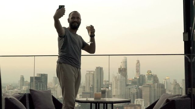 Happy man with glass of wine taking selfie photo with cellphone on roof bar, 4K
