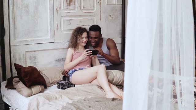 Man and woman in pajamas sitting on the bed. Multiracial couple takes selfie photo, use smartphone and laughing.