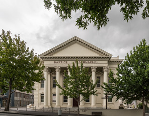 Fototapeta premium Auckland, New Zealand - March 1, 2017: Greek-Roman architecture for beige facade with triangular frieze of Baptist Tabernacle in Queen Street. Green vegetation and line of pillars.