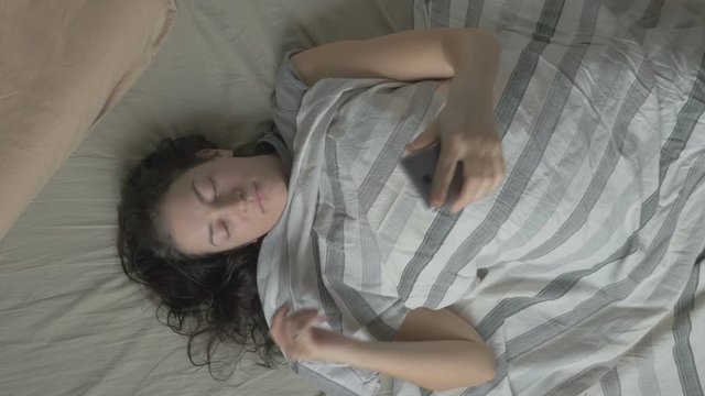 Young woman in bed wakes up in the morning and on the phone . The view from the top