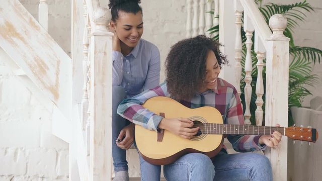 Two brazilian curly girls sistres sitting on stairs and practice to play acoustic guitar. Friends have fun and singing at home