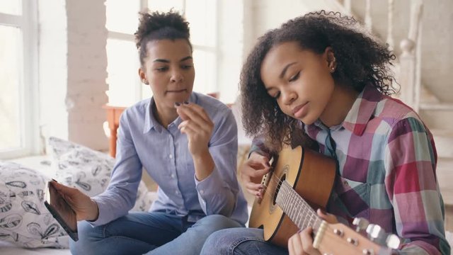 Mixed race young woman with tablet computer sitting on bed teaching her teenage sister to play acoustic guitar at home