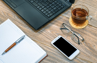 Black Laptop, white mobile phone, tea cup, planner, pen and glasses on wooden table background. education or business concept.