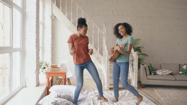 Mixed race young funny girls dancing singing and playing acoustic guitar on a bed. Sisters having fun leisure in bedroom at home