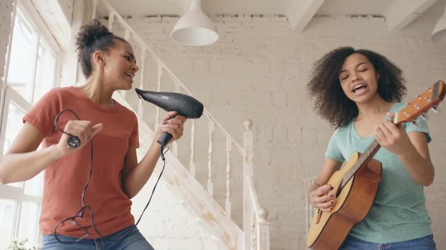 Mixed race young funny girls dance singing with hairdryer and playing acoustic guitar on a bed. Sisters having fun leisure in bedroom at home