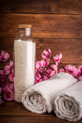 Fototapeta na wymiar Spa or wellness set. White sea salt in Glass bottle, towels and pink flowers on brown wooden background. Selective focus. Place for text.