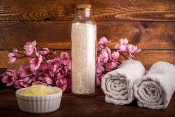 Spa or wellness set. White sea salt in Glass bottle,yellow sea salt in bowl, towels and pink flowers on brown wooden background. Selective focus. Place for text.