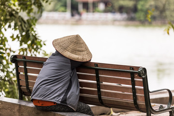 Woman with typical Vietnamese bamboo hat resting on a bench by the lake