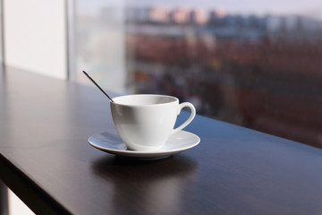 White coffee or tea cup in coffee shop with teaspoon is on the table on the background of a large window overlooking the city. blurred background