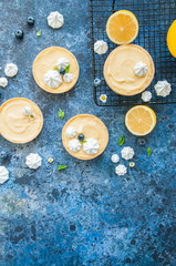 Obraz na płótnie Canvas Individual lemon curd tarts with blueberry jam meringue basil leaves on a blue rusty background. Flat lay and copy space.